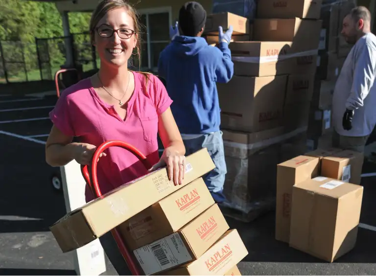 Kaplan delivery person pushing a hand truck stacked with cardboard boxes across the parking lot of an early childcare center