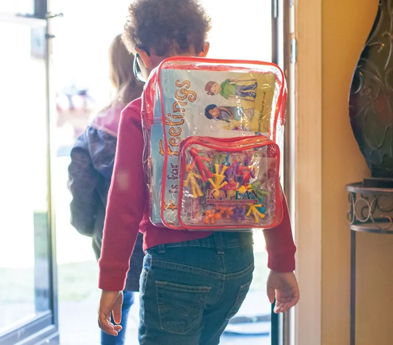 Child walking out a doorway wearing a clear backpack full of educational kit items