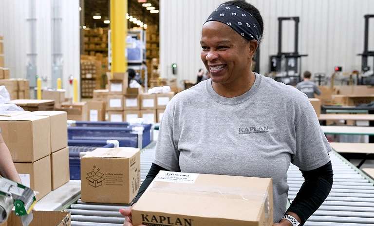 Woman in a warehouse carrying a Kaplan labeled cardboard box and smiling