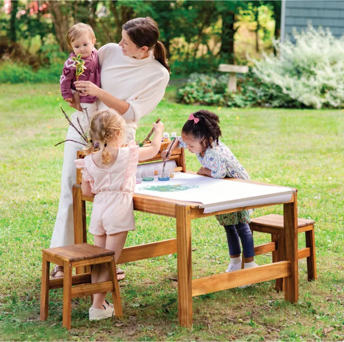 Teacher and children outside painting a large roll of paper on top of a wooden art table