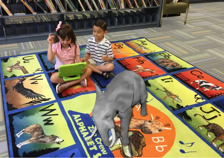 Two children sitting on a classroom rug using the Rugs Alive app on a tablet while a virtual elephant appears in front of them