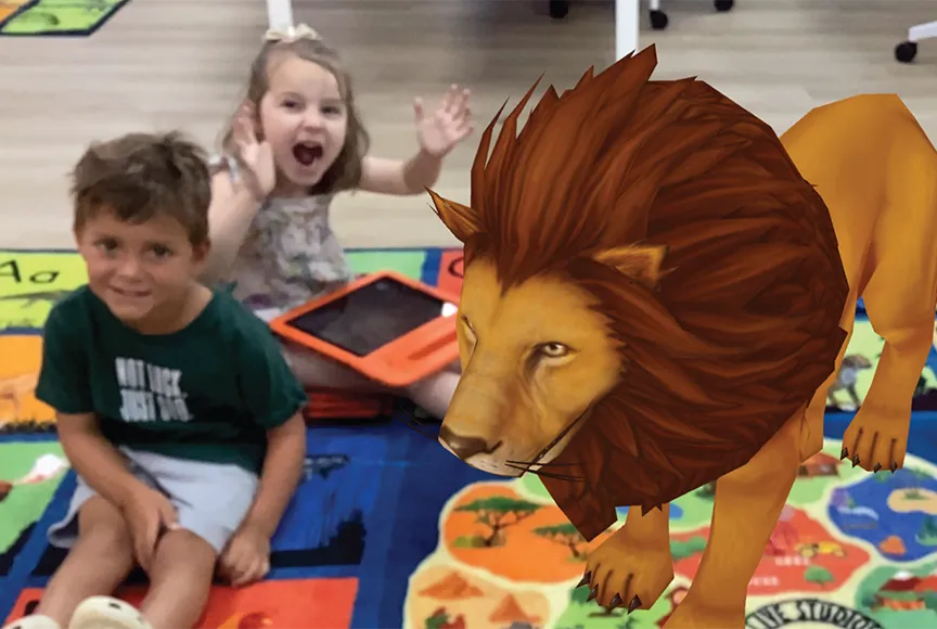 Two children sitting on a classroom rug using the Rugs Alive app on a tablet while a virtual elephant appears in front of them