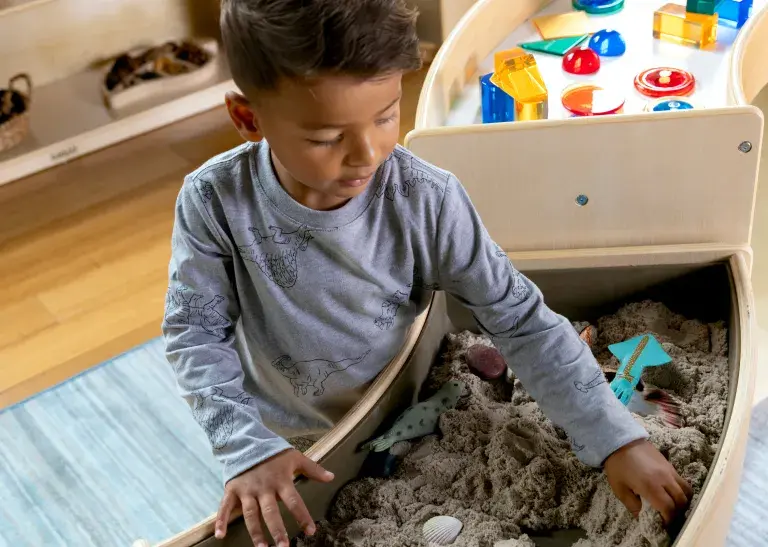 Young child playing with objects in the sand compartment of the Carolina Connections Discovery Table