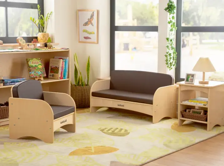Corner of a preschool classroom with wooden furniture showing a shelving unit, couch, and chair on a carpet decorated with a leaf pattern