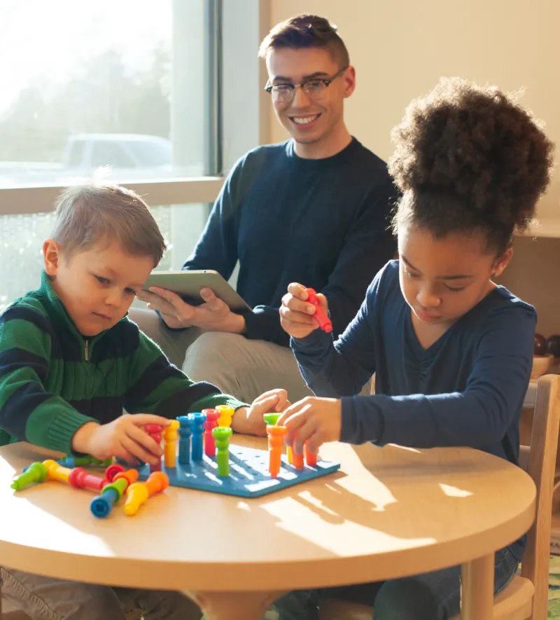 Teacher holding a tablet computer while observing two children engaging in an activity in a classroom