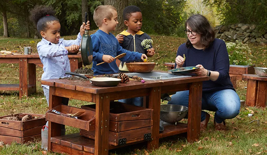 A teacher and children playing outside around the Nature to Play mud kitchen table