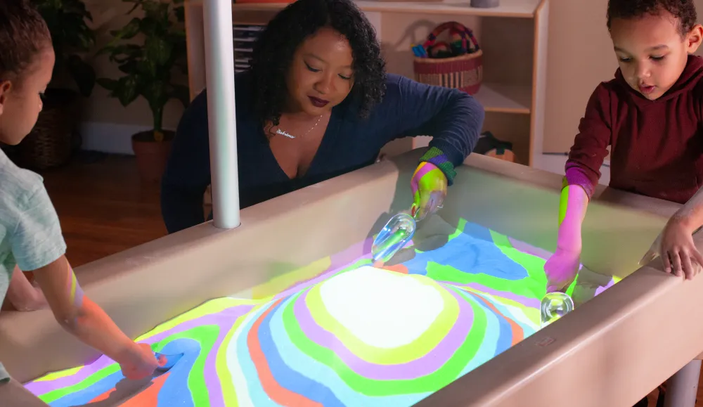 Young children playing in a sand table illuminted in different colors by an overhead projector
