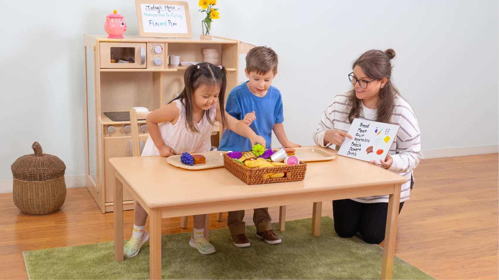 Children playing with toy fruits and vegetables at a table with a woman showing them pictures on a large card